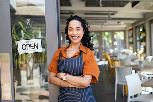Portrait of happy waitress standing at restaurant entrance and looking at camera. Young business woman wearing apron standing with open sign at entrance gate while waiting for clients. Smiling young business owner standing at doorway of her store.