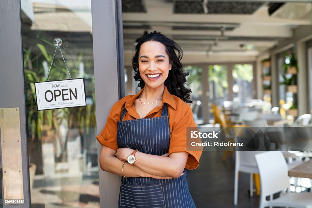 Successful small business owner standing at cafe entrance Portrait of happy waitress standing at restaurant entrance and looking at camera. Young business woman wearing apron standing with open sign at entrance gate while waiting for clients. Smiling young business owner standing at doorway of her store. Owner Stock Photo