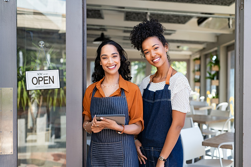 Portrait of smiling young latin woman holding digital tablet with black colleague at cafe entrance door. Two small business owners standing together at cafe entrance while smiling. Happy successful multiethnic small business women wearing apron and standing with open sign at entrance gate of restaurant.