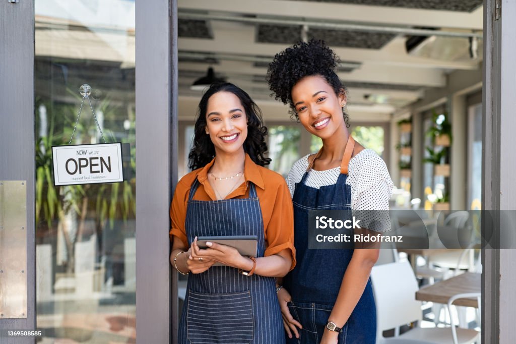 Two successful woman waitress standing at cafe entrance door Portrait of smiling young latin woman holding digital tablet with black colleague at cafe entrance door. Two small business owners standing together at cafe entrance while smiling. Happy successful multiethnic small business women wearing apron and standing with open sign at entrance gate of restaurant. Small Business Stock Photo
