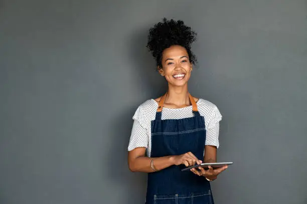 Portrait of beautiful young cafe black waitress using digital tablet on grey background. Happy young black woman in blue jeans apron working on digital tablet while looking at camera. Successful african american cafe owner isolated against gray wall with copy space.