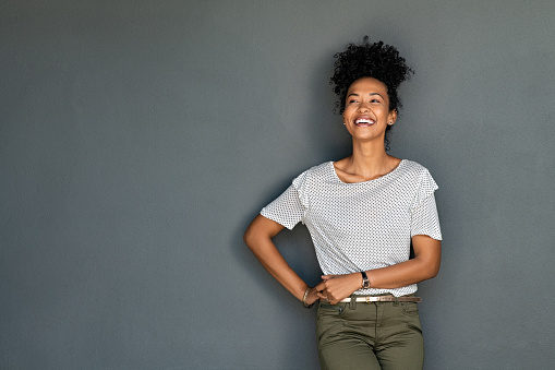 Carefree african woman standing on gray wall with hands on waist while laughing. Confident stylish black girl standing against grey background looking away and smiling. Happy young black woman laughing with copy space.