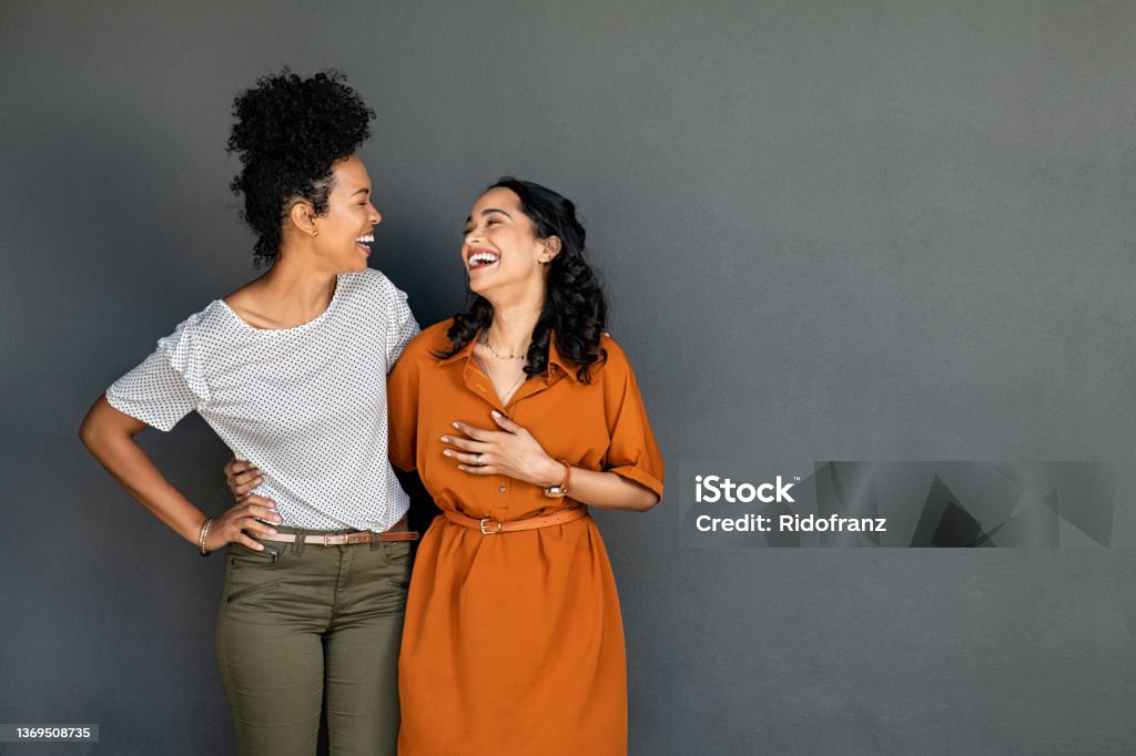 Two women friends embracing and laughing on grey background Happy smiling multiethnic women embracing each other against grey wall with copy space. Happy laughing girls standing on gray background and looking at each other. Carefree african american girl having fun with her latin best friend on grey wall with copy space. Friendship Stock Photo