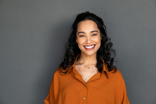 Portrait of latin young woman with pleasant smile isolated on grey wall with copy space. Carefree hispanic woman smiling and looking at camera standing on gray background. Beautiful multiethnic girl in casual clothing laughing against grey wall.