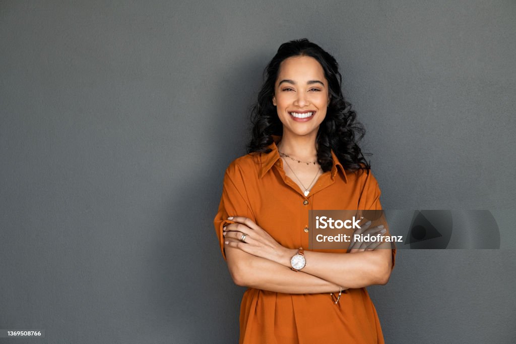 Beautiful successful latin woman smiling Portrait of a young latin woman with pleasant smile and crossed arms isolated on grey wall with copy space. Cheerful hispanic woman on grey background with copy space. Beautiful girl with folded arms looking at camera against grey wall. Women Stock Photo