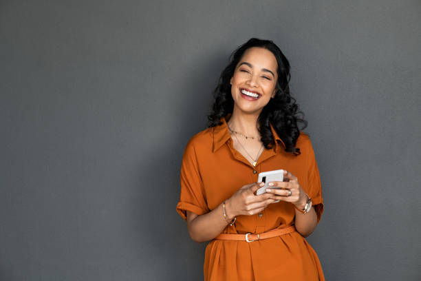 śmiejąca się szczęśliwa uśmiechnięta kobieta wysyła wiadomości na telefon komórkowy na szarej ścianie - happiness cheerful women one person zdjęcia i obrazy z banku zdjęć