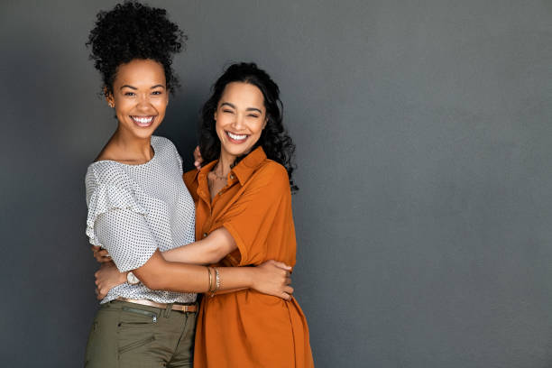 Happy multiethnic girls embracing and laughing on gray wall Portrait of happy women embracing each other against grey wall with copy space. Carefree latin woman hugging her best friend while standing against gray background while looking at camera. Smiling lesbian couple having fun and laughing together. beautiful mexican girls stock pictures, royalty-free photos & images