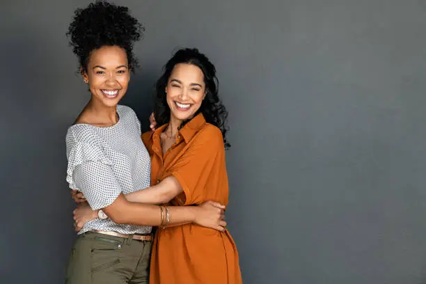 Portrait of happy women embracing each other against grey wall with copy space. Carefree latin woman hugging her best friend while standing against gray background while looking at camera. Smiling lesbian couple having fun and laughing together.