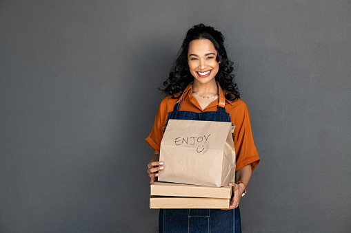 Portrait of smiling cafe waitress holding food box and packet with enjoy message isolated against grey wall. Happy latin woman holding pizza boxes while looking at camera. Young happy mixed race girl holding havana paper packet for take away food order.