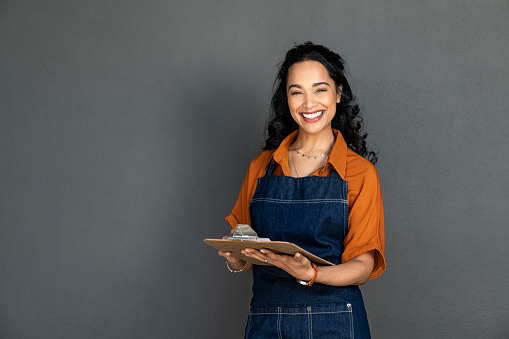 Portrait of smiling young woman in apron holding clipboard and looking at camera. Beautiful latin waitress waiting for customers isolated on grey wall. Successful small business owner standing on grey background with copy space.