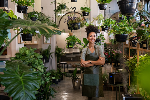 Portrait of african american woman with crossed arms wearing apron standing in botanical store. Smiling young woman in botany store standing between plants looking at camera. Happy small business owner working at flower shop standing surrounded by plants.