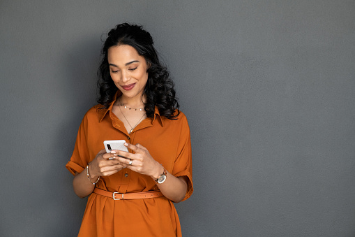 Happy young multiethnic woman writing message on smartphone isolated against grey wall. Beautiful smiling hispanic woman checking mail on mobile phone. Cheerful latin girl chatting with friends by cellphone.