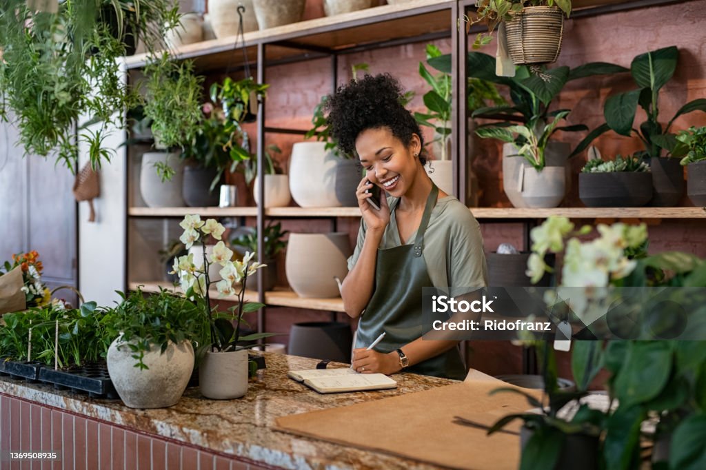 Florist takes an order on the phone at flower shop Smiling woman seller in botanical shop talking on smartphone while writing in diary. African american botanist writing client order in notebook during conversation on mobile phone in store. Young black small business owner wearing apron receive client order for plant and delivery at greenhouse. Small Business Stock Photo