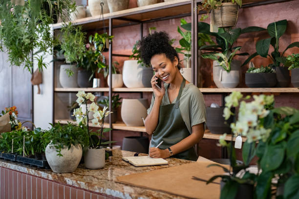 Florist takes an order on the phone at flower shop Smiling woman seller in botanical shop talking on smartphone while writing in diary. African american botanist writing client order in notebook during conversation on mobile phone in store. Young black small business owner wearing apron receive client order for plant and delivery at greenhouse. boutique stock pictures, royalty-free photos & images