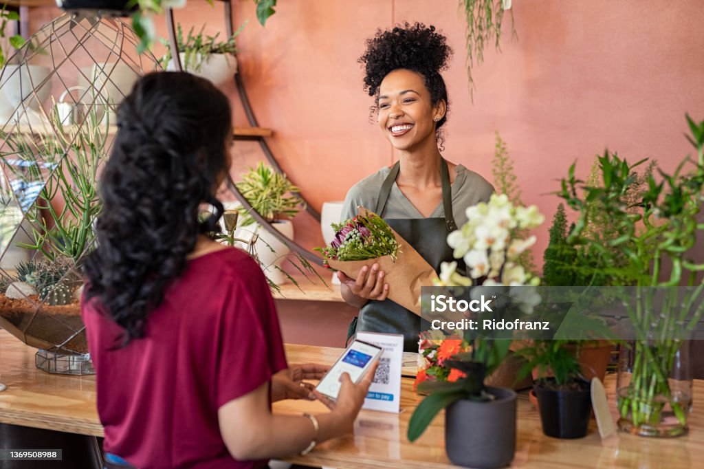 Happy friendly florist giving fresh bouquet to customer Young african florist holding beautiful bouquet of fresh flower while customer making online payment with smartphone while scanning QR code. Black saleswoman wearing apron giving a bunch of flower to customer at botany shop. Young woman making digital payment by phone wallet at flower shop. Small Business Stock Photo