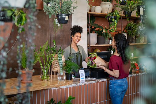 Smiling african woman botanist selling flowers and plants to a customer while standing in flower shop. Happy black young woman entrepreneur standing behind counter wearing apron in plant store selling fresh flowers to client. Young latin girl buy a fresh bouquet from florist.