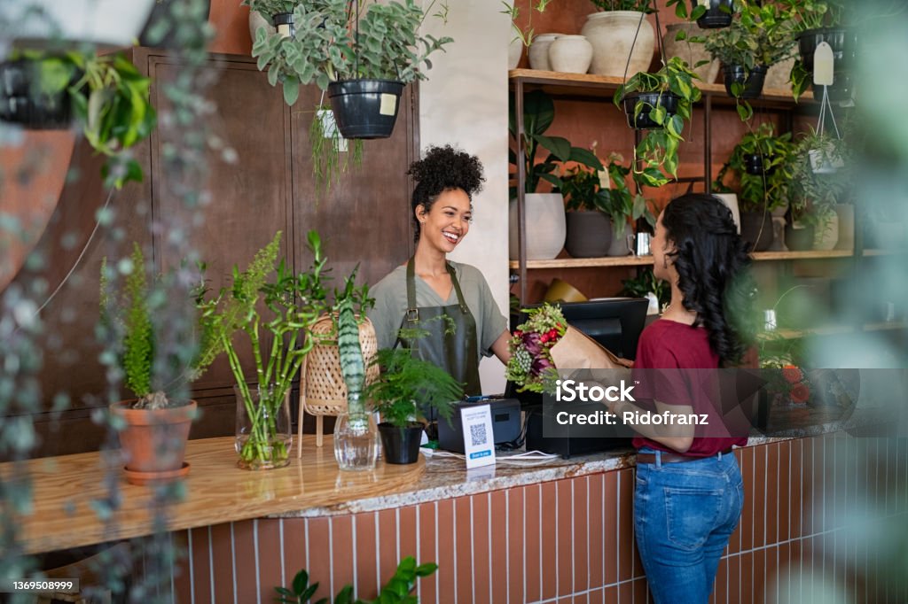 Happy florist selling plants and flower to client Smiling african woman botanist selling flowers and plants to a customer while standing in flower shop. Happy black young woman entrepreneur standing behind counter wearing apron in plant store selling fresh flowers to client. Young latin girl buy a fresh bouquet from florist. Small Business Stock Photo