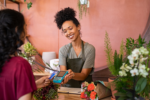 Smiling and friendly florist holding card reader machine at counter with customer paying with credit card. Young african american florist shop assistant holding payment machine while buyer purchase a bunch flower. Woman using bank credit card to make payment by NFC machine.