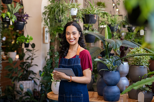 Portrait of smiling young latin woman florist standing and holding digital tablet at floral shop. Successful flower shop owner standing in plant store wearing apron and looking at camera. Happy mixed race entrepreneur in flower shop surrounded by green plants.