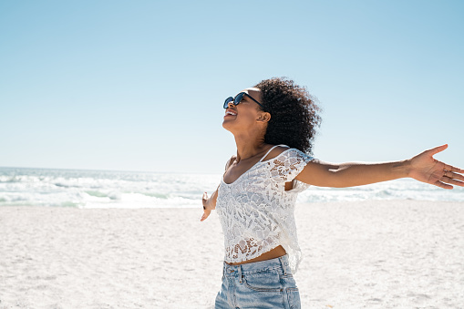 Healthy black woman standing on the beach with her hands outstretched with copy space. Happy young african american woman with open arms at seaside. Freedom girl dancing and daydreaming at beach during summer vacation.
