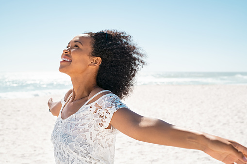 Healthy young african woman with curly hair standing at beach with hands outstretched and looking up. Happy young black woman with open arms feeling the sea breeze with copy space. Beautiful smiling girl with relaxing and enjoy the sun at seaside.