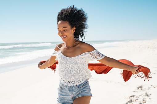 Smiling african american woman running with red scarf enjoying at beach. Beautiful happy black woman holding red fabric at wind and running at seaside. Happy smiling girl holding scarf at wind in summer vacation.
