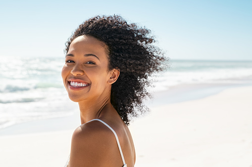 Portrait of young african american woman with curly hair at beach. Smiling black woman walking at the seaside while looking at camera. Happy carefree girl at sea.