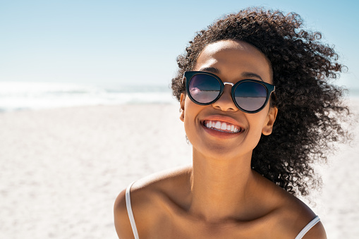 Portrait of smiling black woman wearing fashion sunglasses at the beach with copy space. Pretty african american girl wearing specs at the seaside during summer vacation. Beautiful young woman having fun at sea while looking at camera with copy space.