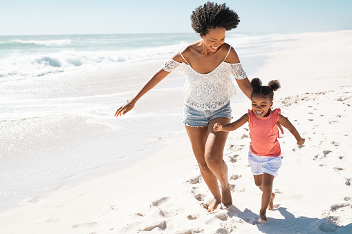 Happy black mom with daughter running on summer beach with copy space. Cheerful african american mother running along seaside with cute little girl with curly hair. Happy older sister having fun with kid at ocean shore.