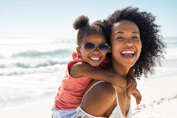 happy young mother giving laughing daughter piggyback ride at beach - laughing beautiful people beauty beautiful imagens e fotografias de stock