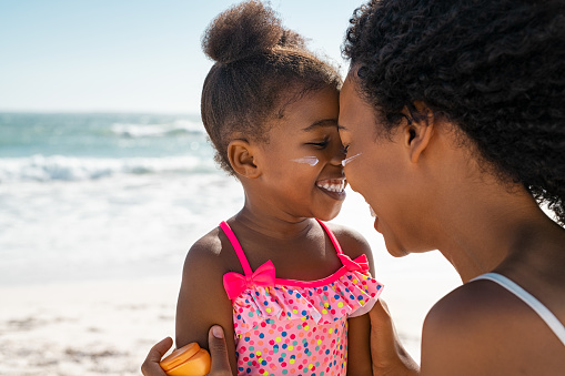 Young black mother embracing daughter with sunscreen on face at beach with copy space. Woman with closed eyes face to face with her cute little girl with sun lotion on face. Funny black female kid with sunblock at seaside with her bigger sister.