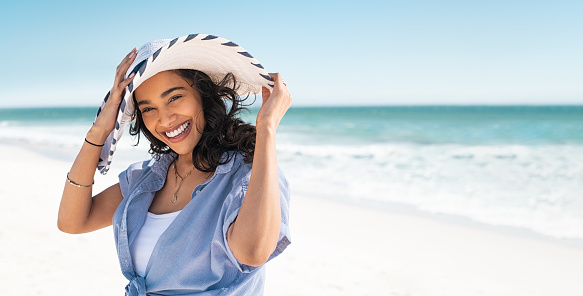 Portrait of stylish latin hispanic woman with white straw hat standing at beach. Young smiling woman on vacation enjoy sea breeze wearing straw hat and looking at camera. Attractive beautiful girl relaxing at seaside.
