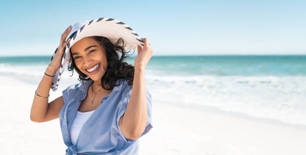 Smiling beautiful latin woman on beach with straw hat at sea Portrait of stylish latin hispanic woman with white straw hat standing at beach. Young smiling woman on vacation enjoy sea breeze wearing straw hat and looking at camera. Attractive beautiful girl relaxing at seaside. beautiful woman summer stock pictures, royalty-free photos & images
