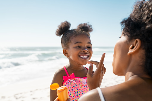 Young mother applying protective sunscreen on daughter nose at beach with copy space. Black woman hand putting sun lotion on female child face. African american cute little girl with sunblock cream at seaside.