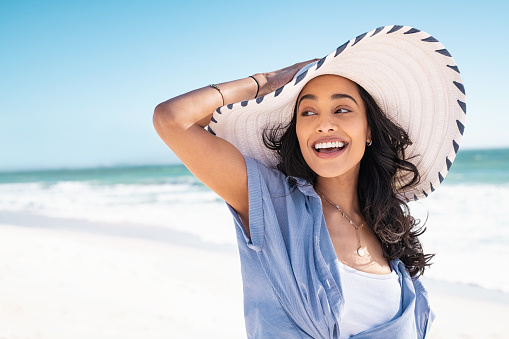 Carefree woman with white and blue straw hat walking at beach. Young smiling latin woman on vacation enjoying sea breeze. Attractive hispanic girl relaxing at beach with the sea behind.