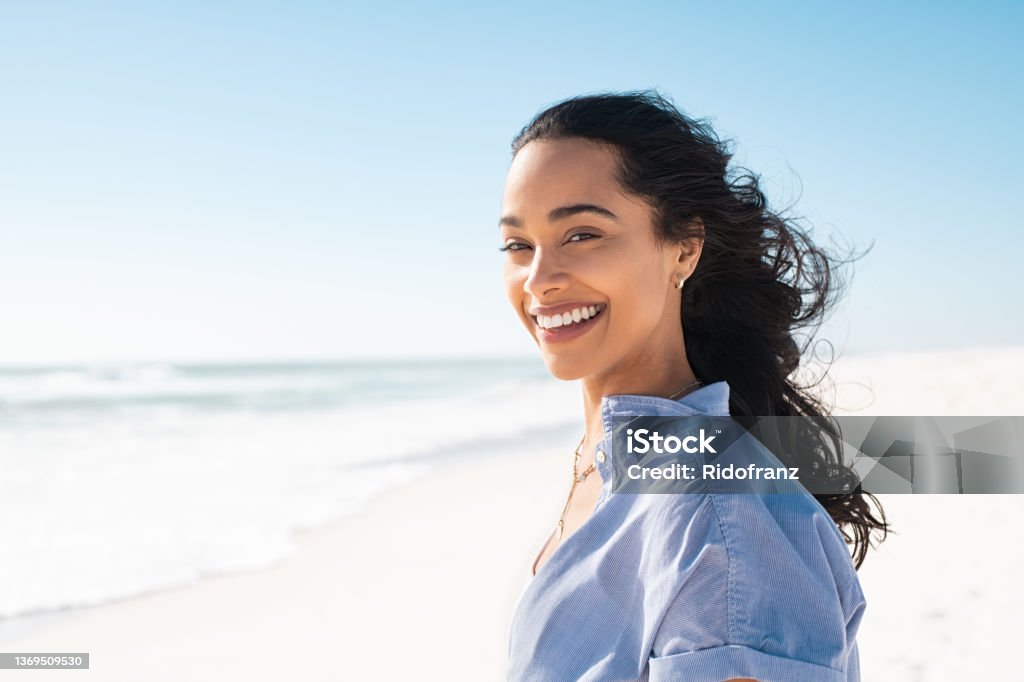 Portrait of natural beauty woman at beach Portrait of young woman at sea looking at camera. Smiling latin hispanic girl standing at the beach with copy space and looking at camera. Happy mixed race girl in casual outfit with wind in her hair. Smiling Stock Photo
