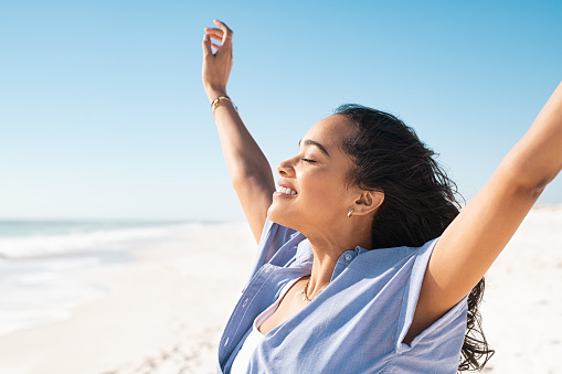 Smiling latin hispanic woman stretching hand and relaxing on beach. Beautiful woman breathing deeply at seaside with eyes closed. Happy woman standing on the beach and enjoy the sun tan with arms outstretched.
