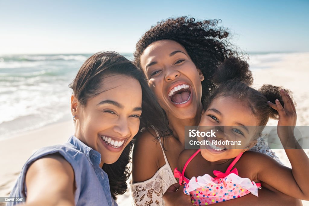 Happy woman friends with child taking selfie at seaside Portrait of smiling young african american woman with child taking selfie at beach with her best friend. Cheerful multiethnic gay couple enjoying at beach with daughter during summer holiday. Happy smiling young mixed race sisters with cute little girl taking selfie over exotic tropical beach. Family Stock Photo