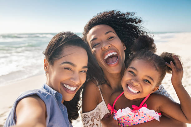 Happy woman friends with child taking selfie at seaside Portrait of smiling young african american woman with child taking selfie at beach with her best friend. Cheerful multiethnic gay couple enjoying at beach with daughter during summer holiday. Happy smiling young mixed race sisters with cute little girl taking selfie over exotic tropical beach. family vacation stock pictures, royalty-free photos & images