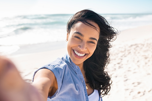Portrait of smiling young woman taking a selfie at beach during sunny day. Cheerful hispanic woman enjoying at beach during holiday. Happy girl taking photo over exotic tropical beach while looking at camera.