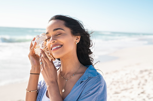 Beautiful latin woman hearing the sound of the sea with a big seashell at beach. Hispanic girl in casual on seacoast with a cockleshell in hands with copy space. Attractive healthy woman on a white sand beach holding a sea shell against her ear and listening the sound of waves during summer vacation.