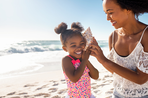 Beautiful young mother holding seashell near happy daughter ear at beach while she listening the sound of the ocean. Cute little female child hearing to cockleshell with mom at tropical beach. Laughing little child with woman listening to a shell at the beach.