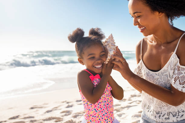 Cute little black girl listening to shell at beach with her mom Beautiful young mother holding seashell near happy daughter ear at beach while she listening the sound of the ocean. Cute little female child hearing to cockleshell with mom at tropical beach. Laughing little child with woman listening to a shell at the beach. seashell stock pictures, royalty-free photos & images