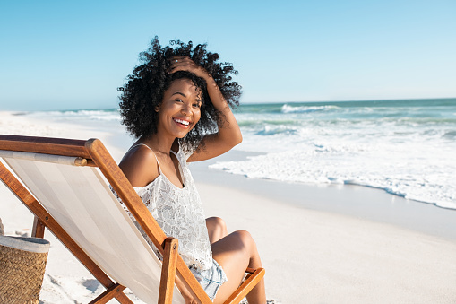 Portrait of happy african american woman relaxing on wooden deck chair at  tropical beach while looking at camera. Smiling black girl enjoying vacation at seaside. Beautiful woman sunbathing and relaxing at sea with copy space.