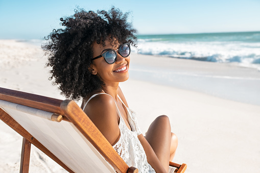 Portrait of happy young black woman relaxing on wooden deck chair at tropical beach while looking at camera wearing spectacles. Smiling african american girl with fashion sunglasses enjoying vacation at sea with copy space. Cheerful young woman sunbathing and relaxing at seaside.