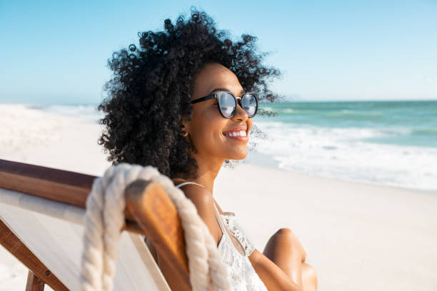 Carefree african woman relaxing on deck chair at tropical beach Portrait of happy young black woman relaxing on deck chair at beach wearing spectacles. Smiling african american girl with sunglasses enjoy vacation at beach. Carefree happy young woman sunbathing and relaxing at sea with copy space. beach holiday stock pictures, royalty-free photos & images