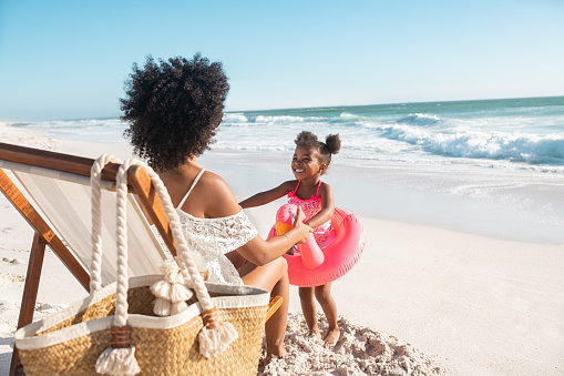 Black cute little girl inside pink float standing on beach with mother while playing together. Rear view of young african american mother sitting on deck chair holding hands of her daughter standing with flamingo inflatable for swim at beach. Young woman with child having fun together at sea.