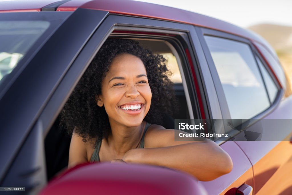 Black young woman looking outside car Beautiful african american woman looking outside car during summer holiday. Young attractive black woman laughing and enjoying road trip on a summer day. Happy girl driving a red car with big grin. Car Stock Photo