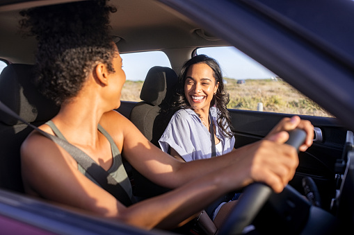 Beautiful latin hispanic woman with best friend enjoying and laughing in car while going on a road trip. Cheerful girl friends enjoying trip and drive in car at sunset. Black woman driving a car with her best firend during summer holiday.
