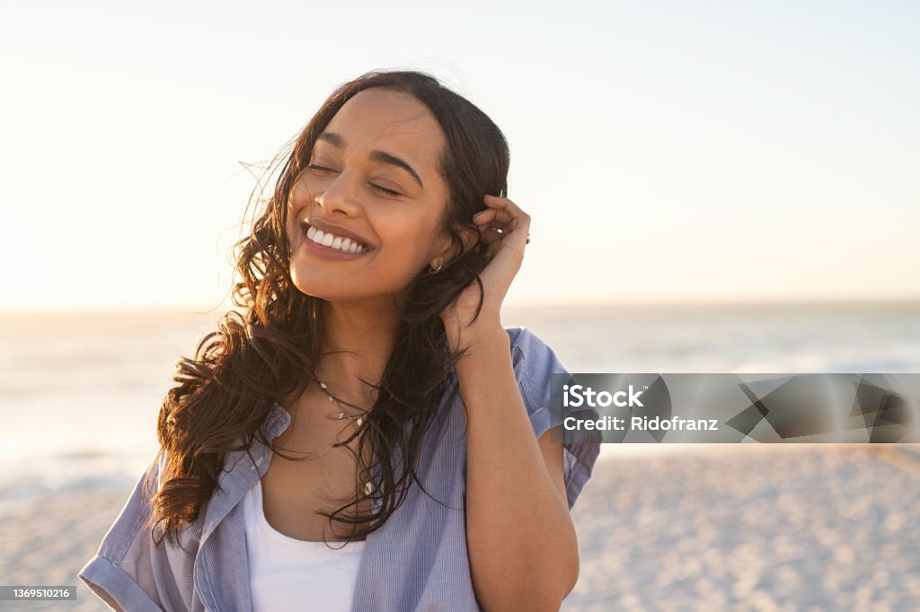 Carefree beautiful latin woman relaxing on beach Smiling latin hispanic woman relaxing on beach with closed eyes at sunset. Beautiful mixed race woman enjoying wind fluttering hair. Charming and calm young woman breathing fresh air at summer beach with copy space. Women Stock Photo